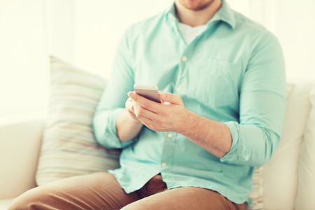 communication, business, home and technology concept - close up of man with smartphone texting message sitting on couch at homeの写真素材