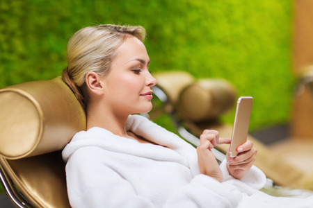 people, beauty, lifestyle, technology and relaxation concept - beautiful young woman in white bath robe with smartphone social networking at spaの写真素材