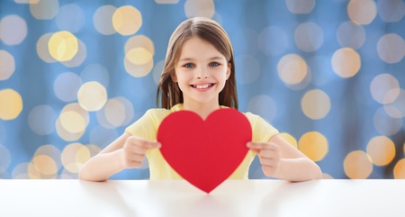 love, charity, holidays, children and people concept - smiling little girl with red heart over blue lights backgroundの写真素材