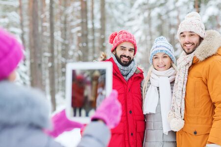 technology, season, friendship and people concept - group of smiling men and women taking picture with tablet pc computer in winter forestの写真素材