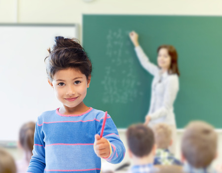 education, elementary school and children concept - happy little student girl with pen over classroom and teacher writing on green blackboard backgroundの写真素材