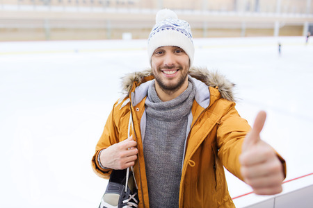 people, sport, gesture and leisure concept - happy young man with ice-skates showing thumbs up on skating rinkの写真素材