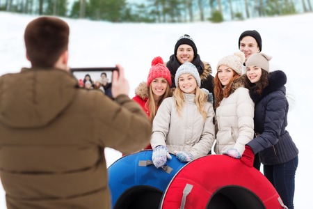 winter, leisure, sport, friendship and people concept - group of smiling friends with snow tubes taking picture by tablet pc computer outdoorsの写真素材