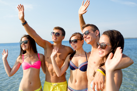 friendship, sea, holidays, gesture and people concept - group of smiling friends wearing swimwear and sunglasses waving hands on beachの写真素材