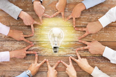 business, people, gesture and inspiration concept - close up of team hands showing victory sign with lighting bulb on table in officeの写真素材