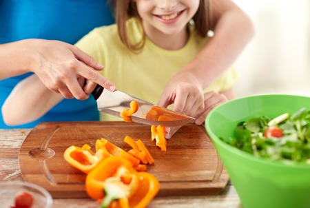 food, family, cooking and people concept - close up of happy girl and mother making salad for dinner and chopping paprika on cutting board with knife in kitchenの写真素材