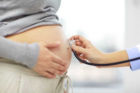 pregnancy, healthcare, people and medicine concept - close up of pregnant woman belly and doctor hand with stethoscope at medical appointment in hospitalの写真素材