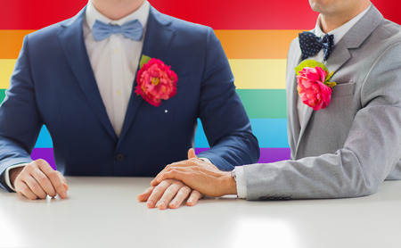 people, homosexuality, same-sex marriage and love concept - close up of happy married male gay couple in suits with buttonholes and bow-ties holding hands on wedding over rainbow flag backgroundの写真素材