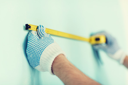 repair, building and home concept - close up of male in gloves measuring wall with tapeの写真素材