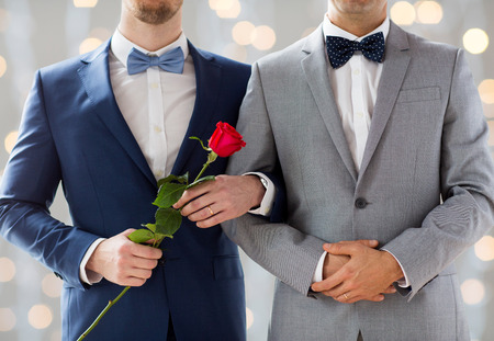 people, homosexuality, same-sex marriage and love concept - close up of happy male gay couple with red rose flower holding hands on wedding  over holidays lights backgroundの写真素材