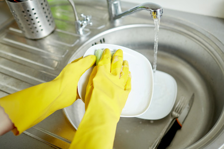 people, housework, washing-up and housekeeping concept - close up of woman hands in protective gloves washing dishes with sponge at home kitchenの写真素材
