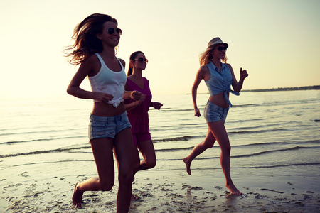 summer vacation, holidays, travel and people concept - group of smiling young women in sunglasses and casual clothes running on beachの写真素材