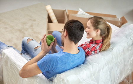 moving, home, repair and people concept - happy couple relaxing and sitting on sofa with cups in new...
