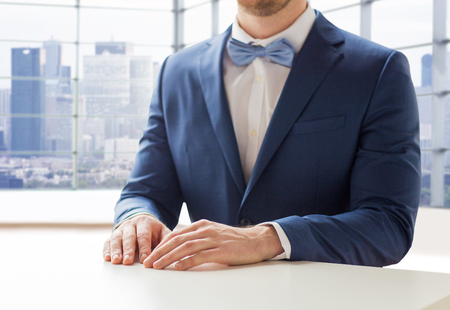 people, fashion, style and wedding concept - close up of best man or groom in suit and bow-tie at table over city office room backgroundの写真素材