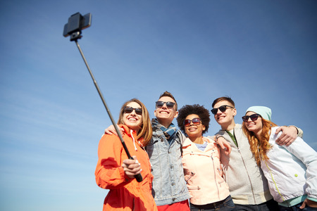 tourism, travel, people, leisure and technology concept - group of smiling teenage friends taking selfie with smartphone and monopod outdoorsの写真素材