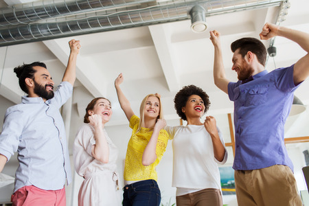 business, triumph, gesture, people and teamwork concept - happy creative team raising hands up and celebrating victory in officeの写真素材