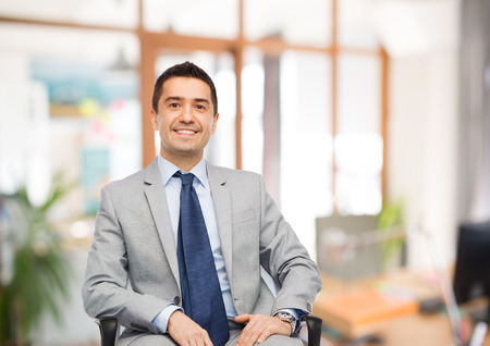 business, people and office concept - happy businessman in suit sitting in chair over office room background