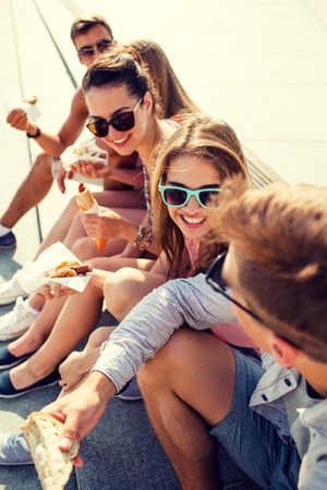 group of smiling friends in sunglasses sitting with food on city squareの写真素材