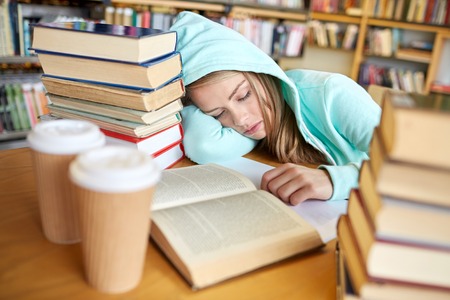 people, education, session, exams and school concept - tired student girl or young woman with books and coffee sleeping in libraryの写真素材
