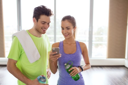 fitness, sport, technology and slimming concept - smiling young woman and personal trainer with smartphone and water bottles in gymの写真素材