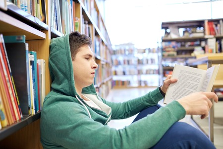 people, knowledge, education, literature and school concept - student boy or young man sitting on floor reading book in libraryの写真素材