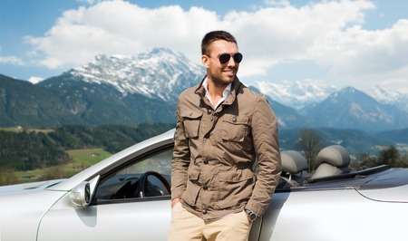 travel, tourism, road trip, transport and people concept - happy man near cabriolet car over mountains backgroundの写真素材
