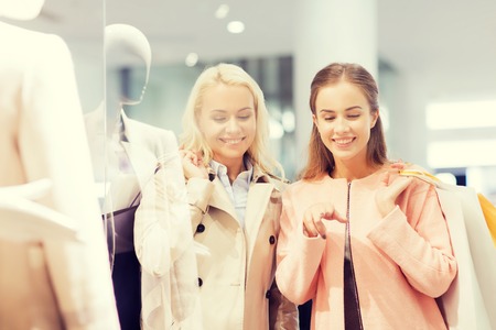 sale, consumerism and people concept - happy young women with shopping bags pointing finger to shop window in mallの写真素材