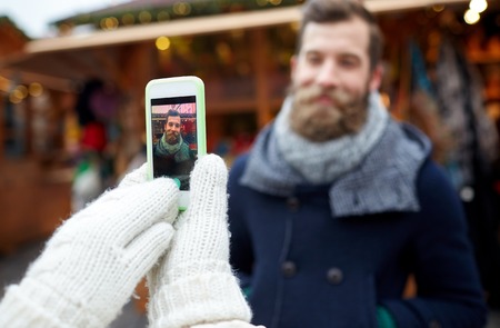 holidays, winter, christmas, technology and people concept - happy couple of tourists in warm clothes taking picture with smartphone in old townの写真素材