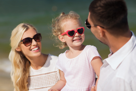 family, vacation, adoption and people concept - happy man, woman and little girl in sunglasses talking on summer beachの写真素材