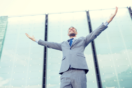 business, people and education concept - young smiling businessman with raised hands over office buildingの写真素材