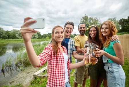 camping, travel, tourism, hike and people concept - happy friends with glass bottles drinking cider or beer and taking selfie by smartphone at campingの写真素材
