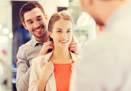 sale, consumerism, presents, holidays and people concept - happy couple trying golden pendant on at jewelry store in mallの写真素材