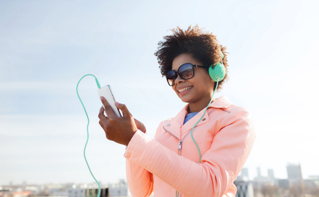 technology, lifestyle and people concept - smiling african american young woman or teenage girl with smartphone and headphones listening to music outdoorsの写真素材