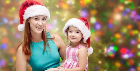 christmas, family, childhood and people concept - happy mother and little girl in santa hats over holidays lights backgroundの写真素材