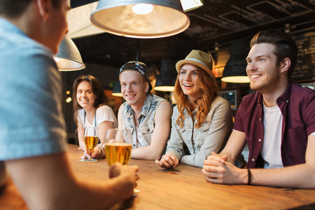 people, leisure, friendship and communication concept - group of happy smiling friends drinking beer and talking at bar or pubの写真素材