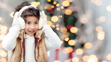 winter, people, childhood and happiness concept - happy little girl wearing earmuffs over christmas tree lights backgroundの写真素材