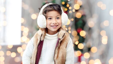winter, people, childhood and happiness concept - happy little girl wearing earmuffs over christmas tree lights backgroundの写真素材