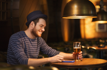 people and technology concept - happy man with smartphone drinking beer and reading message at bar or pubの写真素材