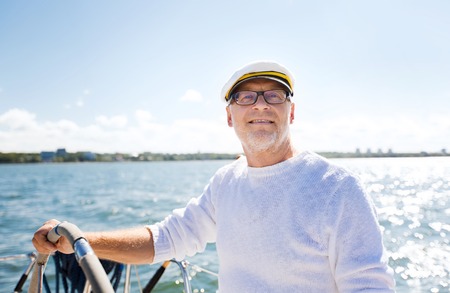 sailing, age, tourism, travel and people concept - happy senior man in captain hat on steering wheel and navigating sail boat or yacht floating in seaの写真素材