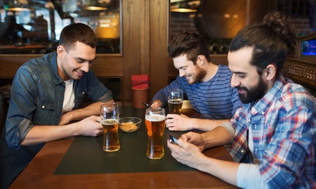 people, men, leisure, friendship and technology concept - male friends with smartphones drinking beer at bar or pubの写真素材