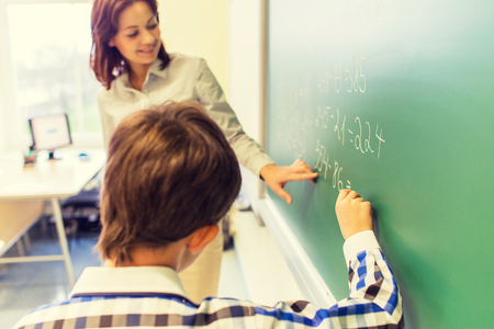 education, elementary school, learning, math and people concept - close up of little schoolboy with teacher writing on chalk board and solving task in classroomの写真素材