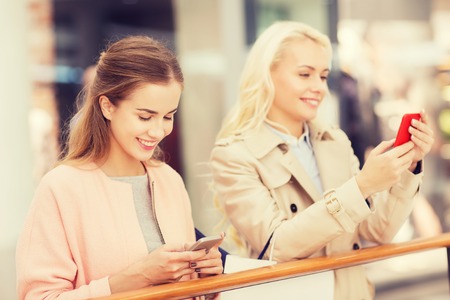 sale, consumerism, technology and people concept - happy young women with smartphones and shopping bags in mallの写真素材