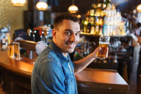 people, drinks, alcohol and leisure concept - happy young man drinking beer at bar or pub