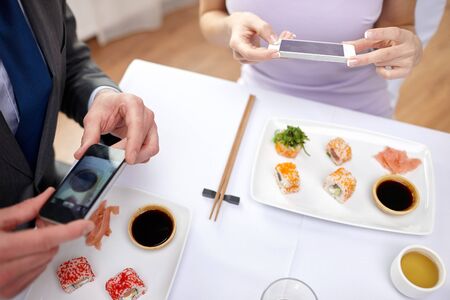 people, leisure, eating, food and technology concept - close up of couple with smartphones taking picture of sushi at restaurantの写真素材