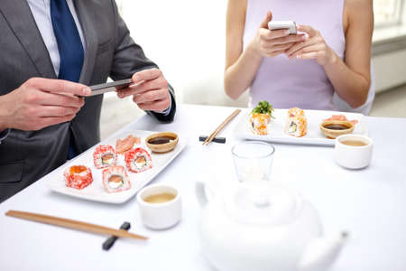 people, leisure, eating, food and technology concept - close up of couple with smartphones taking picture of sushi at restaurantの写真素材
