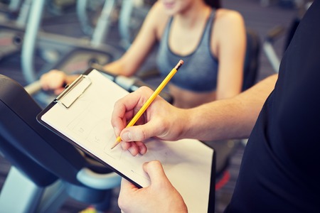 sport, fitness, lifestyle, technology and people concept - close up of trainer hands with clipboard writing and woman working out on exercise bike in gymの写真素材