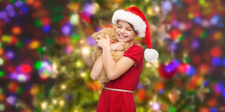 christmas, winter, holidays and childhood concept - smiling girl in santa helper hat with teddy bear over glitter or lights backgroundの写真素材