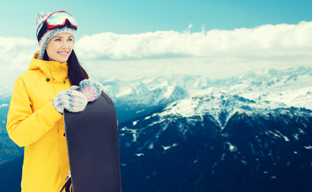 winter, leisure, sport and people concept - happy young woman in ski goggles with snowboard over snowy mountain backgroundの写真素材