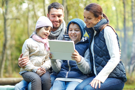 camping, tourism, hike, technology and people concept - happy family sitting on bench with with tablet pc computer at camp in woodsの写真素材