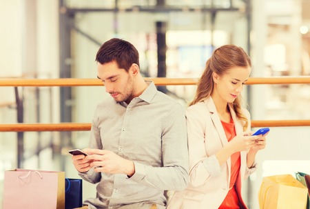 sale, consumerism, technology and people concept - young couple with shopping bags and smartphones in mallの写真素材